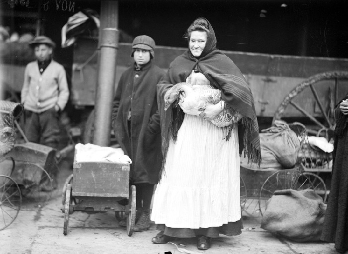 Woman wearing large apron holding a goose in her arms stands with two boys, November 8, 1912