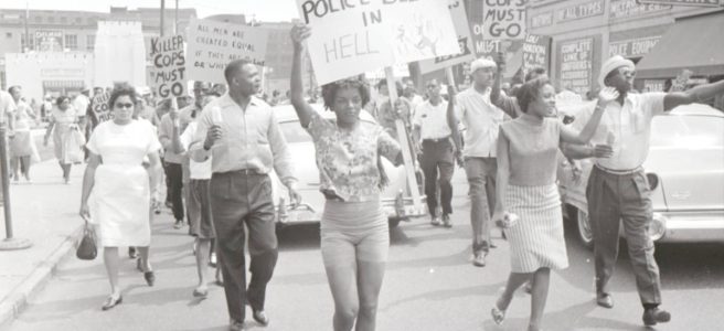 Crowd of people carrying picket signs (one reads, ""killer cops must go,"" another, ""All Gestapo police belong in Hell"") walk down street to protest the fatal shooting of an African American woman by Detroit Police in 1963.